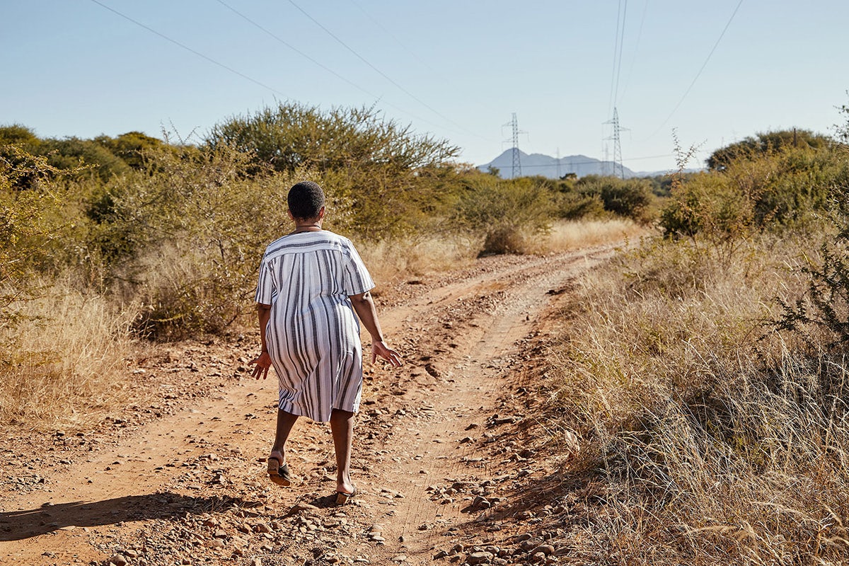 Photo by Thero Makepe of a woman in a dress walking along a dusty track between bushes