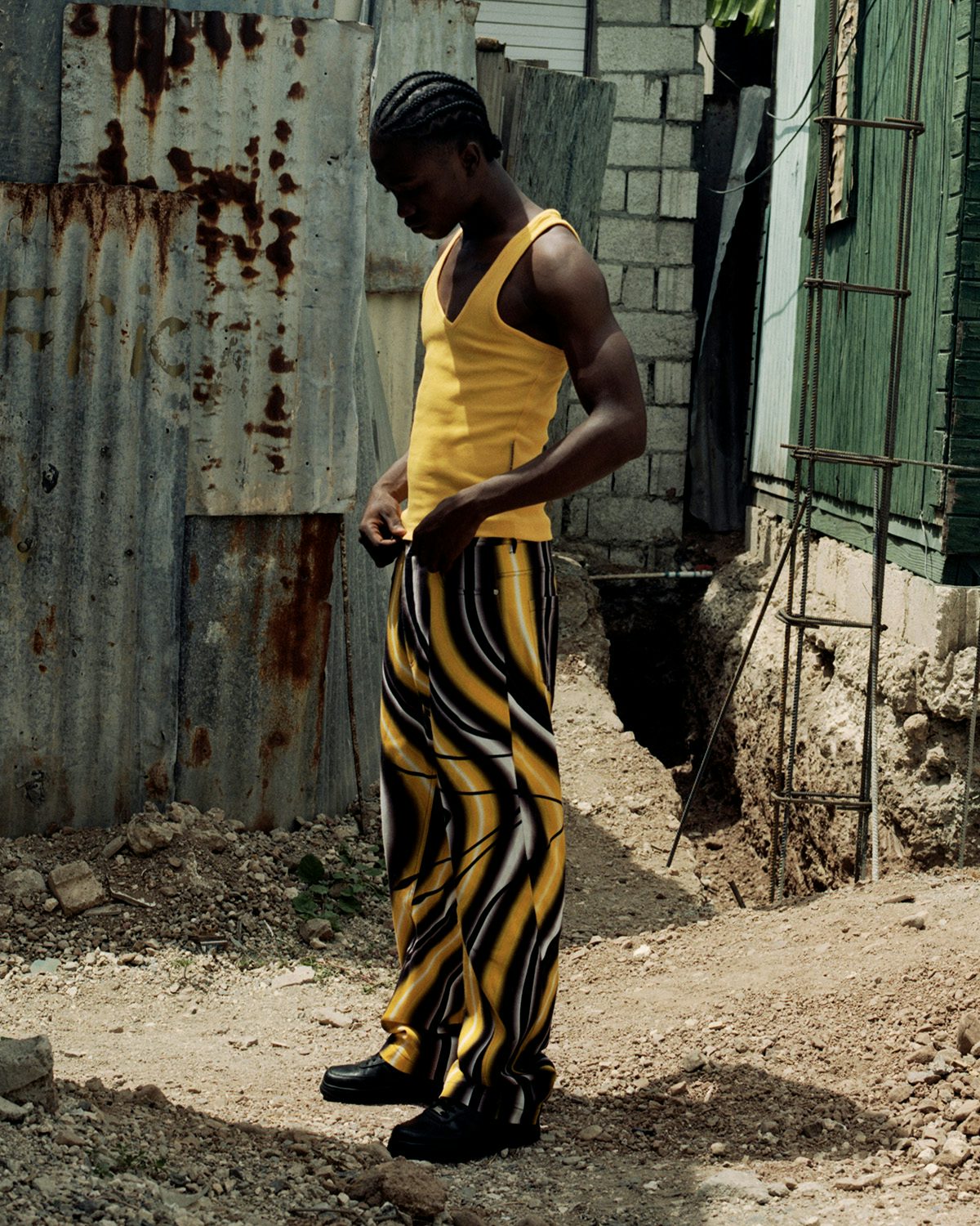 Photo by Kwabena Sekyi Appiah-nti of a person standing in a yard with a corrugated iron building in the background, wearing a yellow vest and black, white and yellow graphic trousers