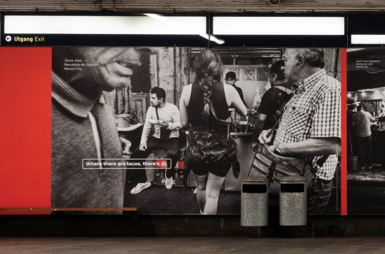 An outdoor ad featuring a black and white photo of a taco stall with a crate of Coca-Cola next to the stall highlighted in red