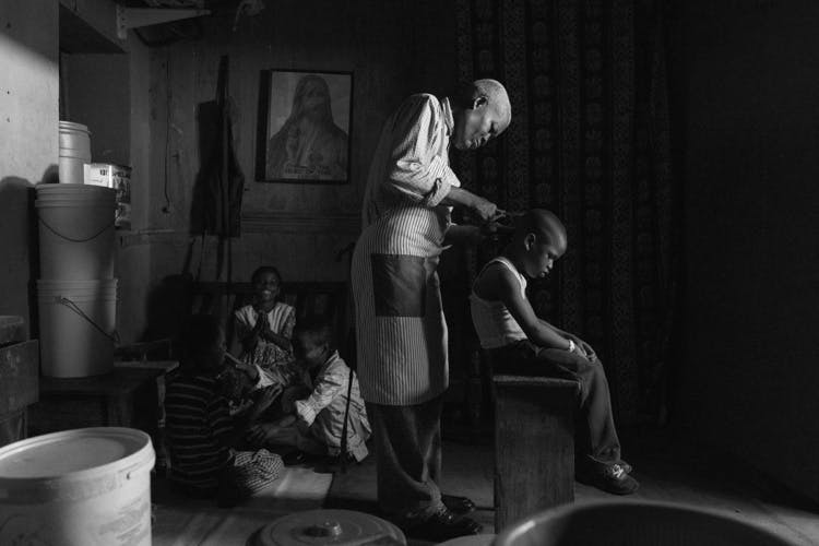 Black and white photo by Carlos Idun-Tawiah showing a barber shaving the hair of a young boy sat on a stool