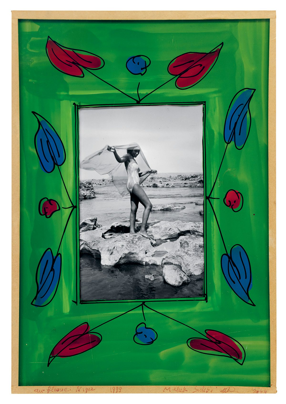 Black and white photo of a person standing along a shoreline waving mesh fabric over their head, which is bordered by an green painted frame with blue and red leaves