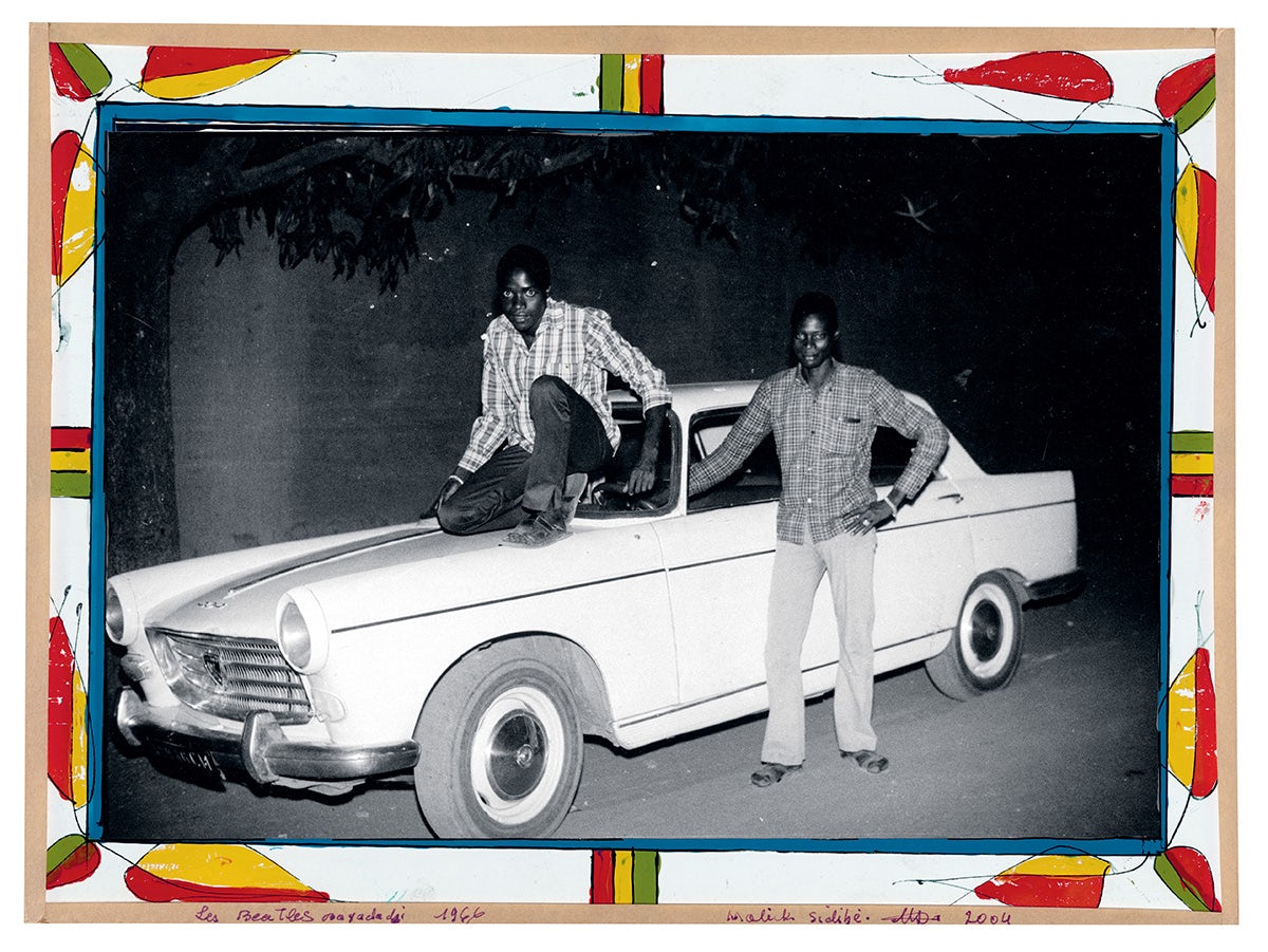 Black and white photo of two people surrounding a white vintage car, which is bordered by a white, yellow and red painted frame