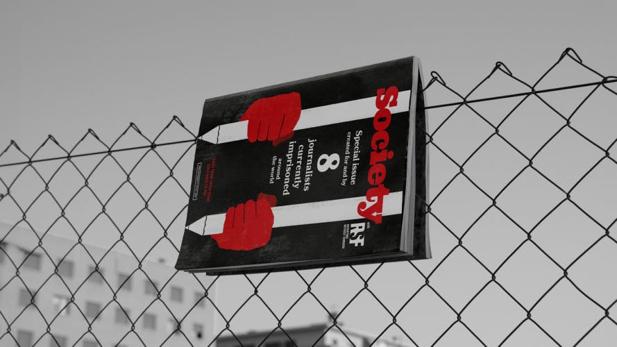 Prison Papers magazine, with an illustrated cover showing hands holding onto prison bars in the shape of pencils. The magazine is shown dangling off a wire fence
