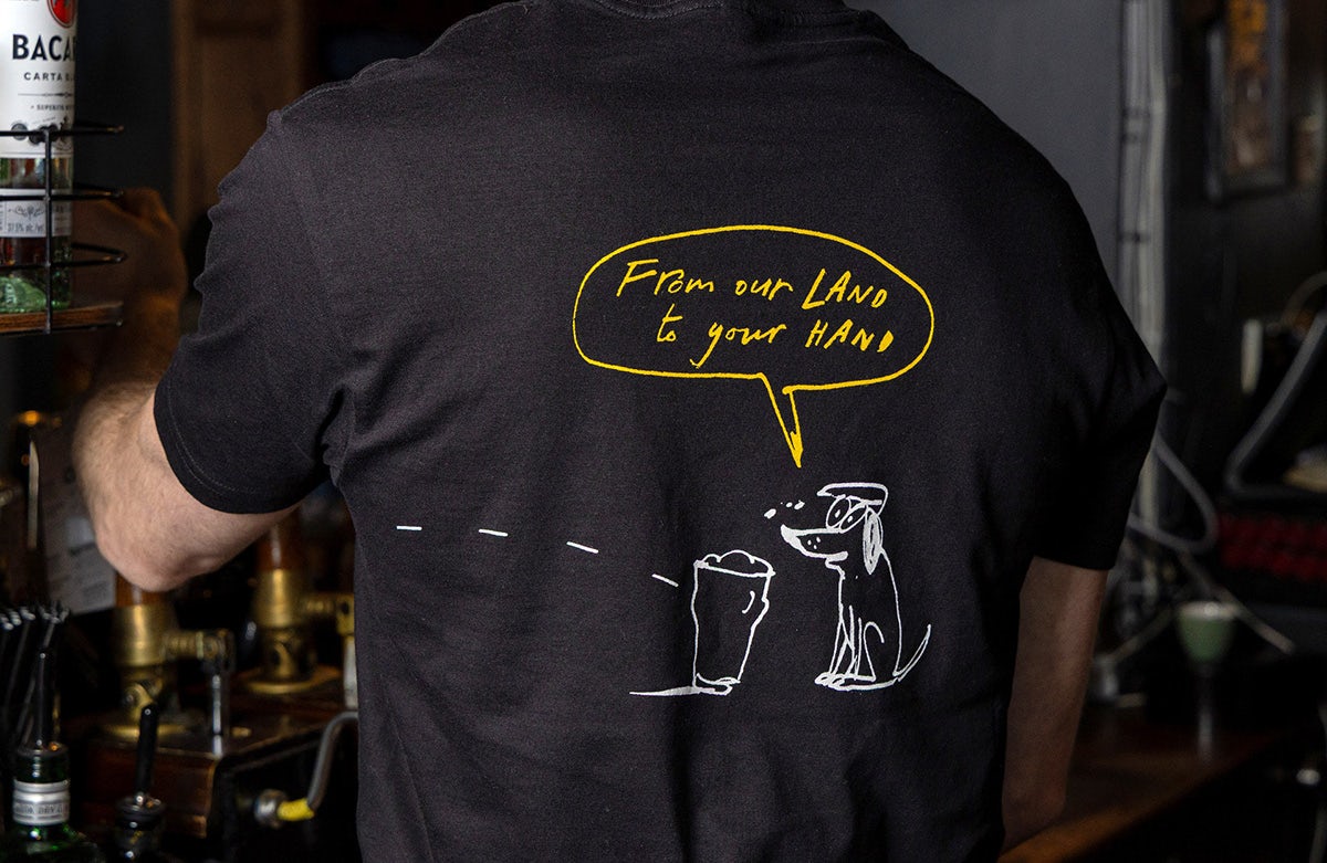 Bartender wearing a black t-shirt featuring a rough illustration of a dog that says 'from our land to your hand' and a pint glass